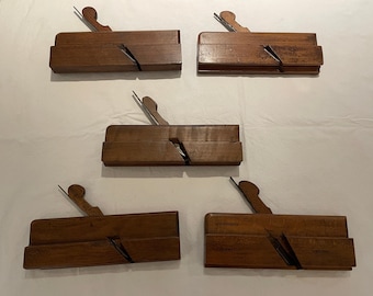 May include: Five antique wooden hand planes, each with a dark brown finish. The planes have a rectangular body with a blade and a handle. They are arranged on a white surface, showcasing their craftsmanship.