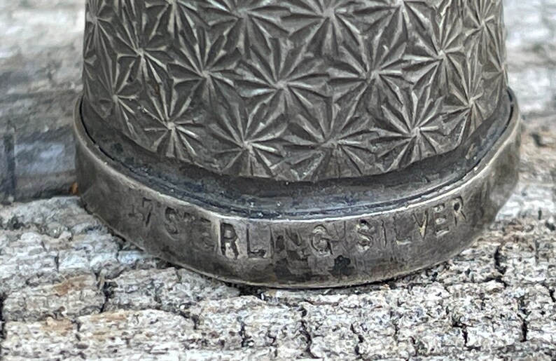 May include: Close-up of a silver thimble with a geometric pattern and the inscription "Sterling Silver" on the bottom.