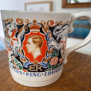 Op de afbeelding: Crèmekleurige keramische mok met een portret van koning Edward. De mok heeft een decoratief ontwerp met een kroon, olifant, hond en bloemmotieven. De woorden "NATION KING EDWARD" zijn in het blauw gedrukt.