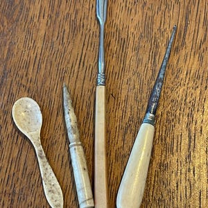 Könnte beinhalten: Eine Sammlung antiker, elfenbeinfarbener Werkzeuge, darunter ein kleiner Löffel, ein spitzes Werkzeug, ein Spatel und ein Messer. Die Werkzeuge haben Metallakzente und sind auf einer Holzoberfläche angeordnet. Der Löffel ist ca. 7.6 cm lang.