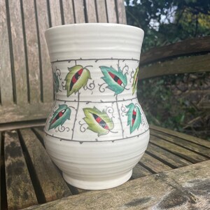 May include: White ceramic vase with a green and red leaf design. The vase has a wide base and a narrow neck.