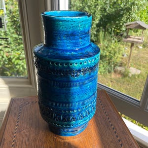 May include: A tall, blue ceramic vase with a textured surface and intricate patterns. The vase features a gradient of blue hues, from a deep teal at the base to a lighter turquoise at the top. The vase is approximately 30 cm tall.