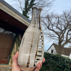 May include: A white ceramic vase with brown glaze and a unique design featuring a series of cut-out holes. The vase has a narrow neck and a wide base.