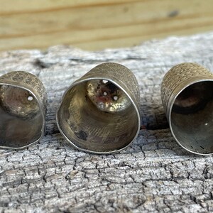 May include: Three silver thimbles of varying sizes, with a textured surface and a circular opening at the top. The thimbles are arranged in a row on a wooden surface.
