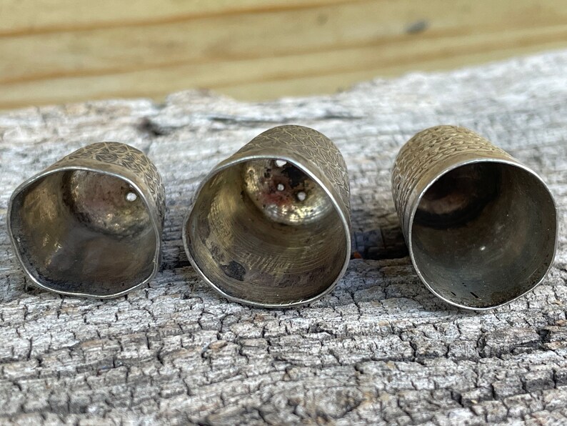 May include: Three silver thimbles of varying sizes, with a textured surface and a circular opening at the top. The thimbles are arranged in a row on a wooden surface.