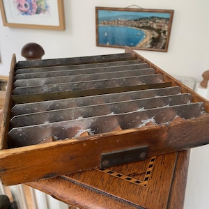 May include: A wooden box with metal dividers, likely vintage. The box is rectangular, made of dark wood, and has a metal plate on the side. The dividers are thin metal strips, creating separate compartments within the box.