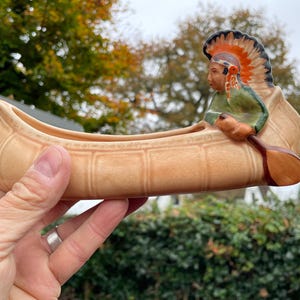May include: A ceramic canoe planter featuring a Native American figure in a headdress paddling. The canoe is a light brown colour with a textured surface. The figure is painted in green, red, and yellow.