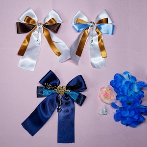 Peut inclure: Trois nœuds pour cheveux faits à la main avec différentes combinaisons de couleurs. Le premier nœud est blanc, marron et or. Le deuxième nœud est bleu, blanc et or. Le troisième nœud est bleu marine avec un charme de serpent doré.