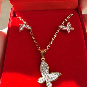 May include: A gold necklace and earrings set with a butterfly pendant and earrings. The butterfly is adorned with white enamel and clear rhinestones.