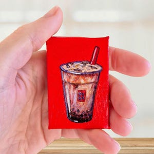 May include: A small rectangular canvas painting featuring a detailed illustration of a brown bubble tea drink with ice cubes and a red straw, set against a solid bright red background. The drink cup displays a small red square logo.