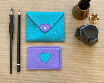 May include: Two felt envelopes with a heart design. The larger envelope is turquoise blue with a purple heart. The smaller envelope is purple with a turquoise blue heart.