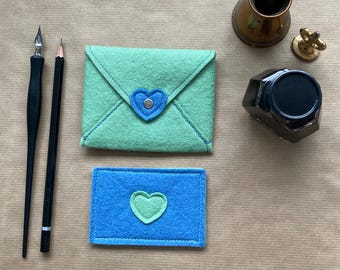 May include: A stationery set featuring a green felt envelope with a blue heart closure and a blue felt card with a green heart. A pencil, pen, inkwell, and dip pen are also present, all arranged on a brown surface.