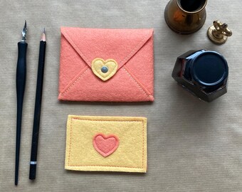 May include: Two felt envelopes with a heart design. The larger envelope is peach colored with a yellow heart. The smaller envelope is yellow with a peach heart.