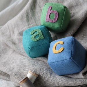 May include: Three felt alphabet blocks, one green with a pink letter "b", one teal with a green letter "a", and one blue with a yellow letter "c". The blocks are on a gray and white fabric.