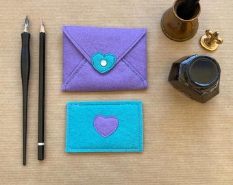 May include: Two felt envelopes with a heart design. The larger envelope is purple with a teal heart. The smaller envelope is teal with a purple heart.
