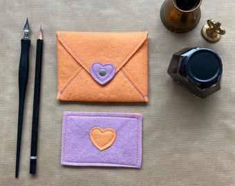 May include: An orange felt envelope with a purple heart-shaped clasp and a matching purple and orange felt card. A black pen, pencil, inkwell, and a brass ink holder are also present. The items are arranged on a brown surface.