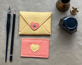 May include: Two felt envelopes with heart-shaped cutouts. The larger envelope is yellow with a pink heart, and the smaller envelope is pink with a yellow heart.