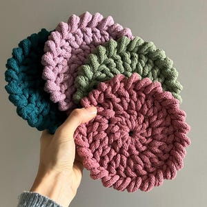 Peut inclure: Un ensemble de quatre dessous de verre circulaires crochetés à la main en fil épais et texturé. Les dessous de verre présentent un motif en spirale tressé et sont disponibles en quatre couleurs : bleu sarcelle, lavande, vert sauge et vieux rose.