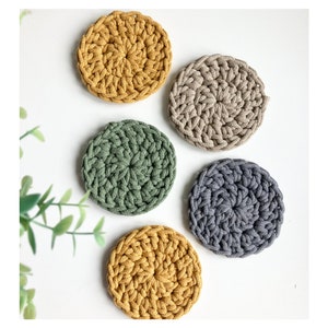 Puede incluir: Cinco posavasos de ganchillo en diferentes colores. Dos son de color amarillo mostaza, uno es verde, uno es gris y uno es beige.