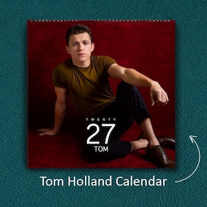 K&ouml;nnte beinhalten: Ein Wandkalender mit einem Foto von Tom Holland, der ein olivgr&uuml;nes T-Shirt und eine dunkle Hose vor einem tiefroten Hintergrund tr&auml;gt. Das Kalenderblatt zeigt den Text TWENTY 27 TOM in wei&szlig;er Schrift mittig &uuml;ber dem Bild platziert.