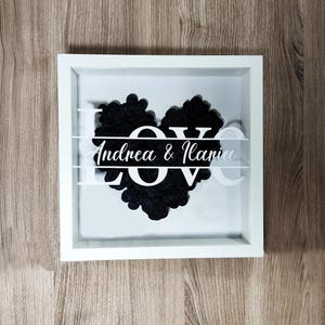 Può includere: Scatola ombra incorniciata in bianco con un cuore di fiori di carta nero che scrive "LOVE" con i nomi "Andrea & Ilaria" sotto il cuore.