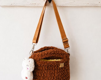 May include: A brown crochet handbag with a thick, textured yarn construction, featuring a long tan adjustable strap with metal hardware. A small white plush cat charm hangs from the side, and a yellow fabric lining is visible inside the opening.