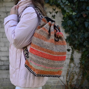 May include: A woman wearing a pink puffer jacket and white pants is carrying a large, colorful woven backpack with a drawstring closure. The backpack has a geometric pattern in shades of orange, red, black, and white.
