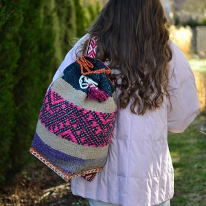 May include: A woman wearing a pink puffer jacket and white pants is carrying a colorful woven bag with a tassel. The bag has a geometric pattern in shades of pink, purple, black, and brown.