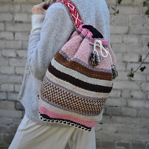 May include: A woven drawstring backpack in shades of pink, brown, and white, with a patterned strap. The bag features a mix of textures and a decorative tassel. The strap has a red and white geometric design.