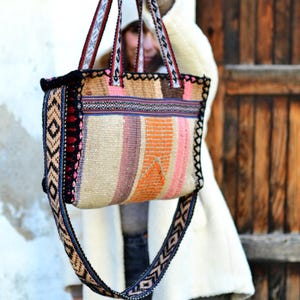 Peut inclure: Un sac fourre-tout coloré avec un motif tissé. Le sac présente des rayures verticales dans les tons orange, rose et beige. Les sangles et les bords sont ornés de garnitures à motifs. Le sac est présenté devant une porte en bois.