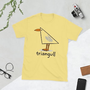 May include: A light yellow short-sleeve t-shirt featuring a graphic of a bird shaped like a triangle with an orange beak and feet, grey scribbled wing detail, and the text "triangull" printed in black lettering below the bird illustration.