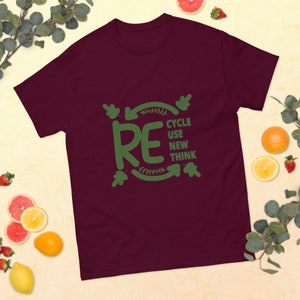 Könnte beinhalten: Ein kastanienbraunes Kurzarm-T-Shirt mit einem grünen Grafikdesign, das den Text RE CYCLE USE NEW THINK innerhalb von kreisförmigen Pfeilen und Blattakzenten zeigt. Das Design ist mittig auf der Brust des T-Shirts mit Rundhalsausschnitt platziert.