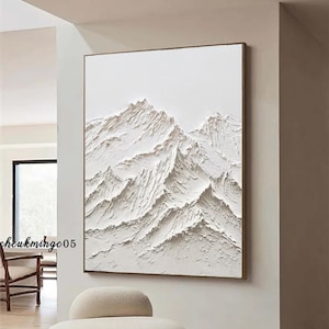 Pode incluir: Uma pintura em tela branca texturizada que representa uma cordilheira de montanhas sobre um fundo branco. A pintura está pendurada em uma parede em uma sala com uma cadeira branca em primeiro plano.