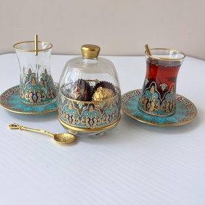 Könnte beinhalten: Dekoratives Teeservice mit zwei klaren Gläsern mit Goldrändern, verzierten blau-goldenen Designs und kleinen goldenen Löffeln. Zwischen den Gläsern befindet sich eine Glas-Bonbonniere mit goldenem Deckel und passendem Design.