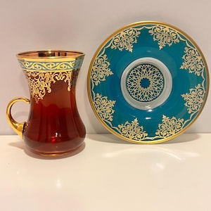 Peut inclure: Un ensemble de deux tasses à thé turques avec des bordures dorées et des motifs complexes. Les tasses sont en verre transparent et présentent un motif bleu sarcelle et blanc. La soucoupe est bleu sarcelle avec des bordures dorées et des motifs complexes.