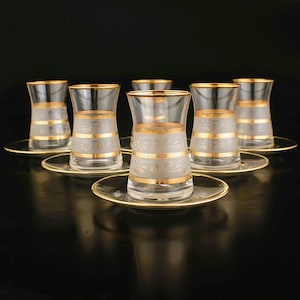 Peut inclure: Ensemble de six tasses à thé en verre transparent avec des bordures dorées et des motifs gravés. Les tasses sont disposées sur des soucoupes assorties.