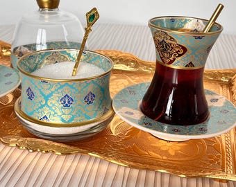 May include: A gold tray with a blue and gold patterned tea set. The set includes a teapot, two teacups, two saucers, a sugar bowl, and a sugar spoon. The teacups are filled with dark tea.
