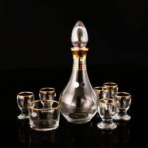 May include: A set of clear glass decanter and shot glasses with gold rims. The decanter has a clear glass stopper with a gold top.