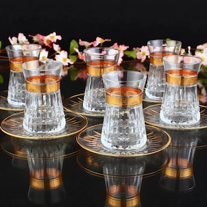 Peut inclure: Ensemble de six tasses à thé en verre transparent avec une bordure dorée et des soucoupes assorties. Les tasses ont un motif cannelé et sont disposées sur une surface noire.
