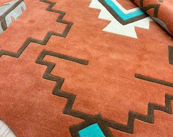 Puede incluir: Una alfombra de color terracota con patrones geométricos en zigzag de color marrón oscuro, un cuadrado de color turquesa brillante y motivos de media luna y plumas en gris claro. La alfombra tiene una textura gruesa y está parcialmente doblada.