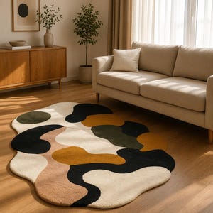 May include: A modern area rug with an abstract design. The rug features a cream-colored base with large, irregular shapes in black, olive green, mustard yellow, and tan. The rug is placed on a hardwood floor in a living room setting.