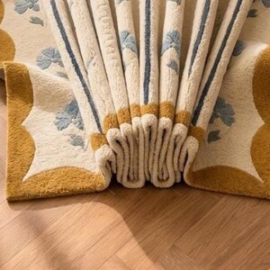 May include: A cream-colored area rug featuring a repeating pattern of light blue floral motifs and dark blue vertical stripes. The rug has a thick, textured pile and a wide mustard yellow border with scalloped edges, folded to show its depth.