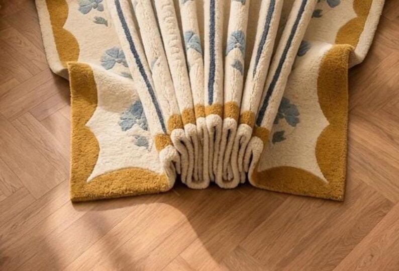 May include: A cream-colored area rug featuring a repeating pattern of light blue floral motifs and dark blue vertical stripes. The rug has a thick, textured pile and a wide mustard yellow border with scalloped edges, folded to show its depth.