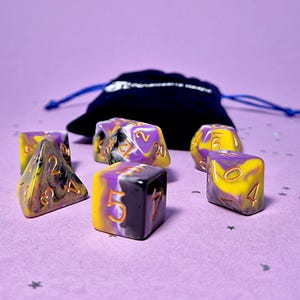 Könnte beinhalten: Ein Set aus sieben Würfeln mit mehreren Seiten in Gelb, Lila und Schwarz mit Marmormuster. Die Würfel haben goldfarbene Zahlen und liegen auf einer lila Oberfläche, im Hintergrund ein schwarzer Beutel. Für Rollenspiele.