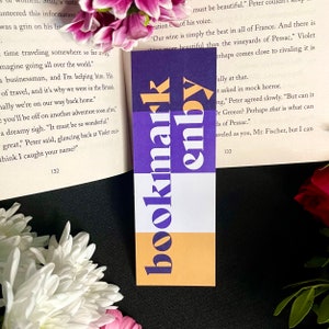 Könnte beinhalten: Ein violettes, weißes und gelbes Lesezeichen mit den Wörtern "bookmark" und "enby" in einer fetten, serifenlosen Schrift.