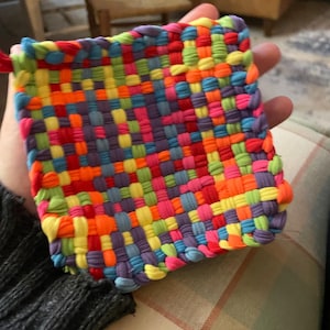 Op de afbeelding: Een kleurrijke, geweven pannenlap in vierkante vorm, gemaakt van stoffen stroken. Het ontwerp heeft een patchwork van kleuren: rood, oranje, geel, groen, blauw en paars. Er is een kleine lus bevestigd om op te hangen. Handgemaakte, getextureerde uitstraling.