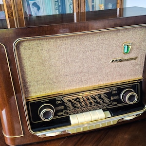 Op de afbeelding: Een vintage tafelradio met een gepolijste donkerbruine houten behuizing en gouden accenten. De voorzijde is voorzien van een beige stoffen luidsprekerrooster, een centrale afstemknop met zwarte en gouden markeringen en vijf witte rechthoekige drukknoppen.