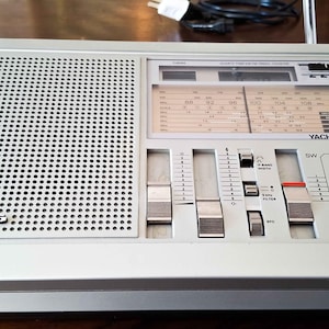 Puede incluir: Una radio Grundig Yacht Boy 700 plateada con una rejilla de altavoz, diales de sintonización y una pantalla. La radio tiene una antena retráctil y varios botones para diferentes bandas de onda. La radio está sobre una superficie de madera.