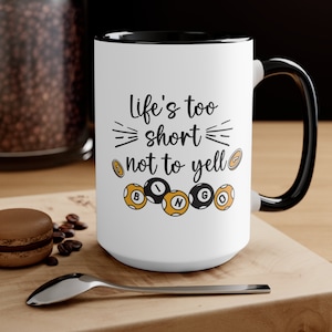 May include: White ceramic mug with a black rim and handle. The mug has a black and white graphic that says "Life's too short not to yell BINGO".