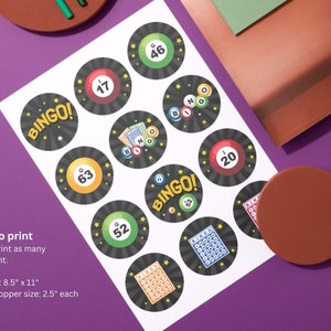 May include: A sheet of printable bingo cupcake toppers. Each black circle features a bingo ball number or the word "BINGO!" in gold. The paper size is 8.5" x 11", and each topper is 2.5" in diameter. The background is purple.